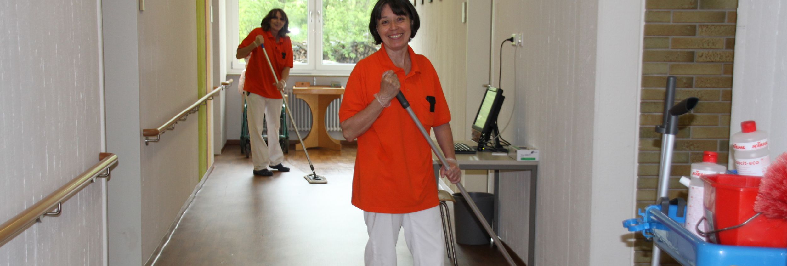 Zwei Reinigungskräfte in orange Polos wischen einen langen Flur